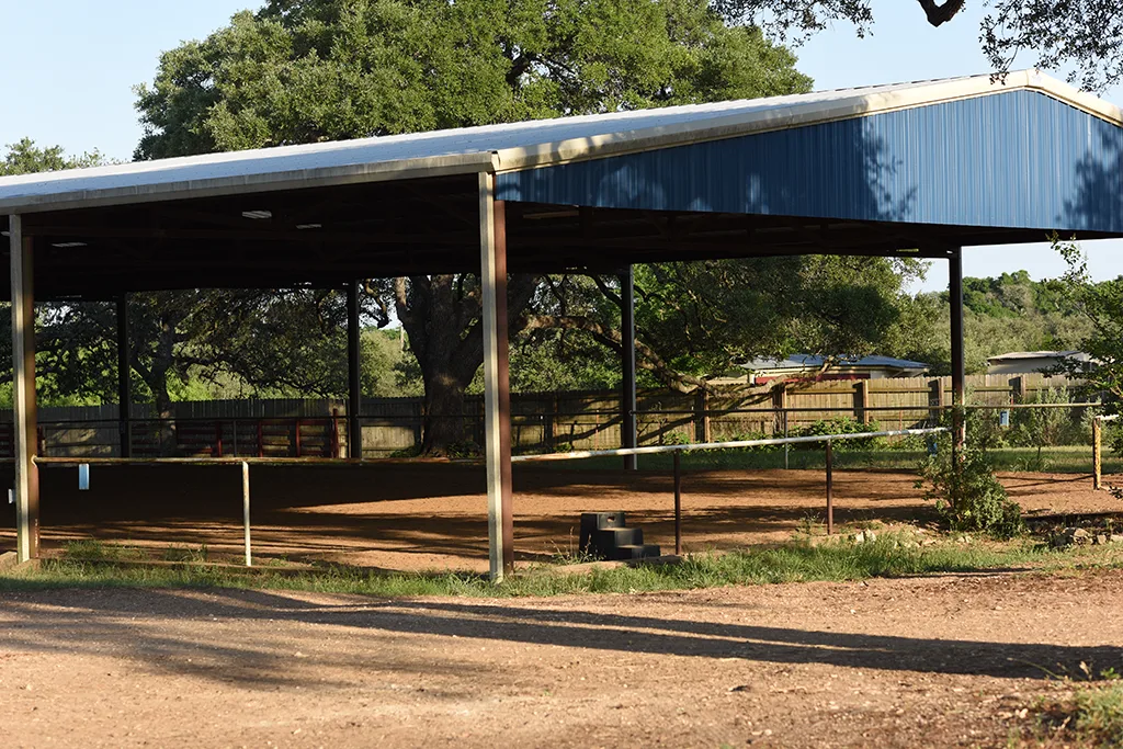 La arena cubierta abierta de Blue Moon Farm con cercas tubulares, superficie de tierra y grandes árboles de sombra alrededor.